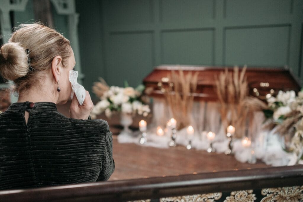 Within the poignant church setting, the image showcases a casket surrounded by an abundance of flowers, while a grieving woman, clutching flowers, sits in sorrow, exemplifying coping with the loss of a family member.