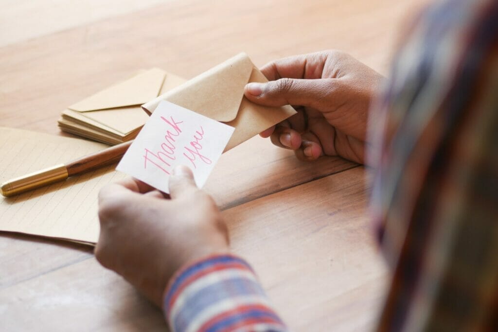 A touching scene of appreciation: A teacher seated in a classroom, focused on opening a small card placed on a table in front of her. The card reveals a heartwarming 'Thank you note,' symbolizing the gratitude from students.