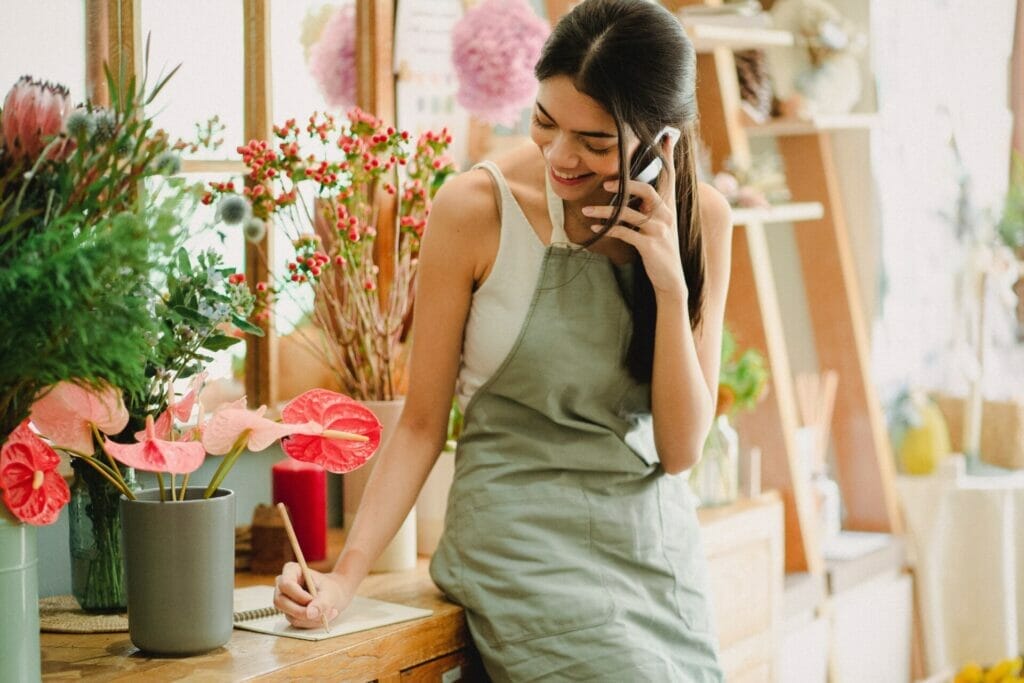 In this charming flower shop scene, a beautiful girl stands, joyfully engaging in small talk over the phone while simultaneously jotting down notes, radiating a warm smile.