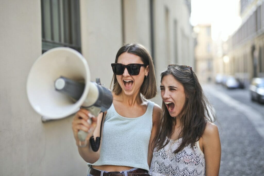 In this lively snapshot, two young girls are immersed in an animated conversation, both radiating infectious smiles. Their enthusiasm is palpable as they joyfully shout into a speaker, sharing a moment that embodies 'living the dream.' Their genuine connection and spirited interaction capture the essence of living life to the fullest, truly embodying the art of responding when someone says 'living the dream'."