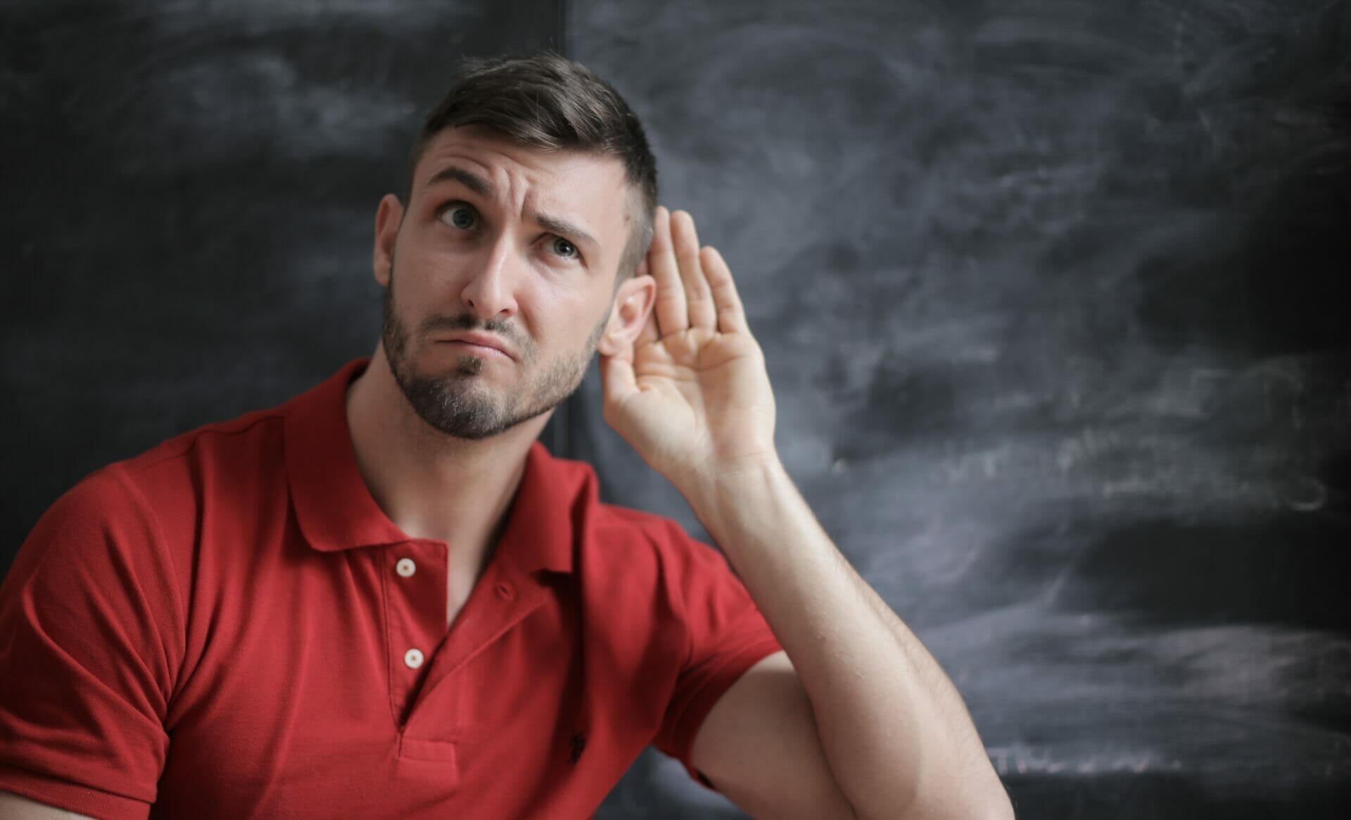 In a room, a man wearing a bright red T-shirt stands, visibly perplexed as he raises his hand to his ear in a signal of struggle, illustrating the difficulties and challenges that individuals encounter in the realm of active listening.