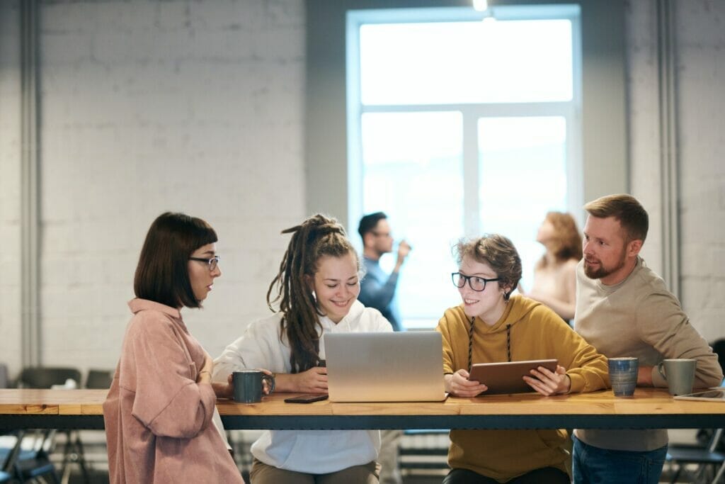 In this scene, three young girls and a man are standing in a room, with two of the girls focused on their laptop, actively discussing what is challenging about active listening. The other man is smiling and attentively observing the conversation.