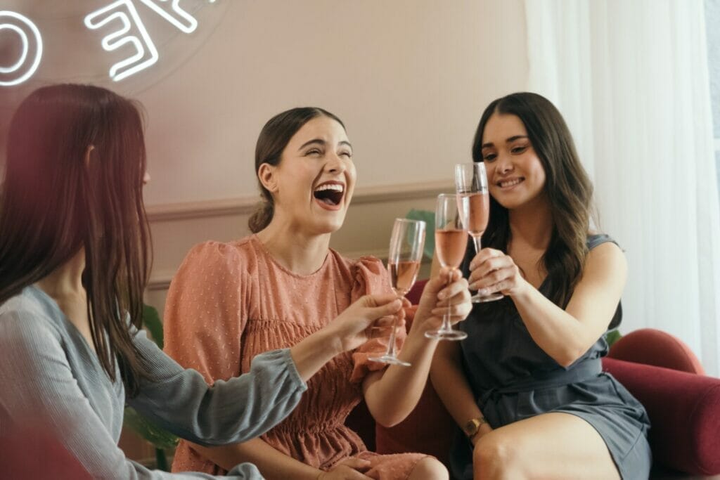 Three young girls sitting in a hotel room, celebrating with cheerful toasts, and raising their drinks in joyous bachelorette party festivities. This image perfectly aligns with the theme of 'Bachelorette Party Captions for Sister.' bachelorette party captions and quotes for sister
